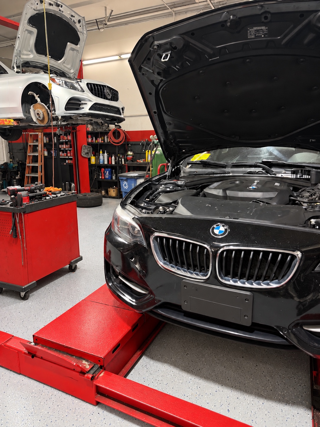 Black BMW with its hood open and a white Mercedes on a lift inside the Legacy Auto Repair workshop